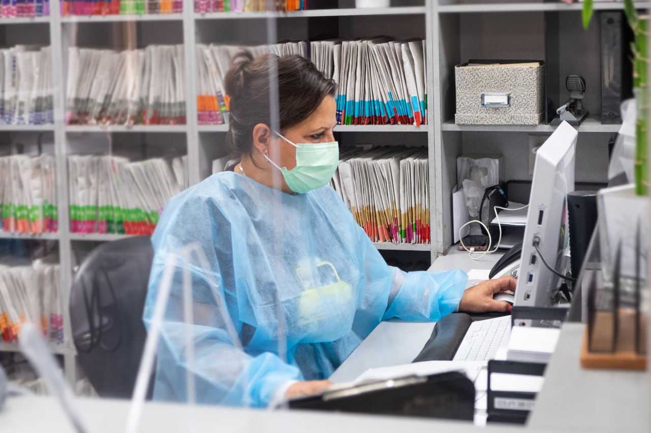 在Covid-19大流行期间，牙科诊所的西班牙裔接待员戴着口罩和防护服. 她正在电脑前工作，后台是病人的档案.